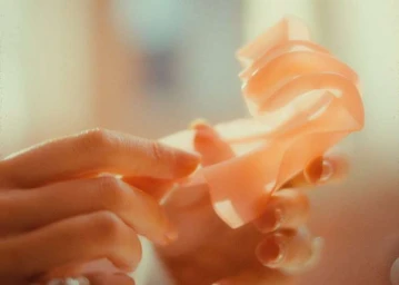 Close-up of hands gently shaping a translucent peach-coloured ribbon under soft warm lighting by Mathieu Richer Mamousse