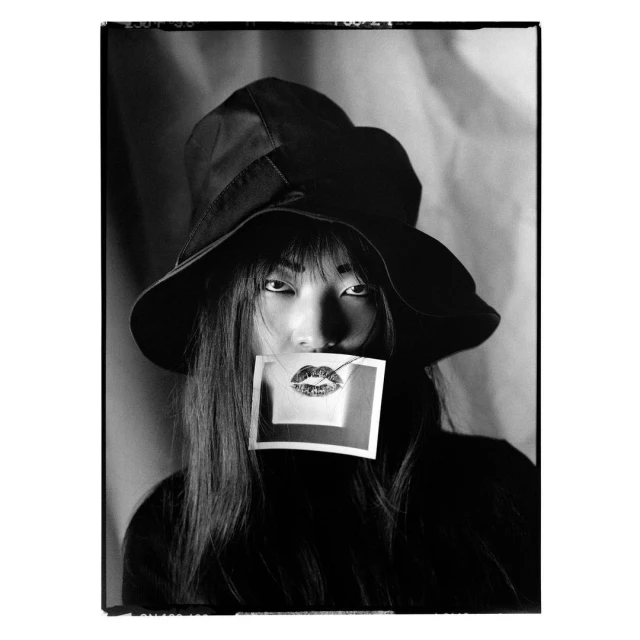 Portrait of Asian woman wearing black hat and holding image of red lipstick kiss against wavy white background in black and white.