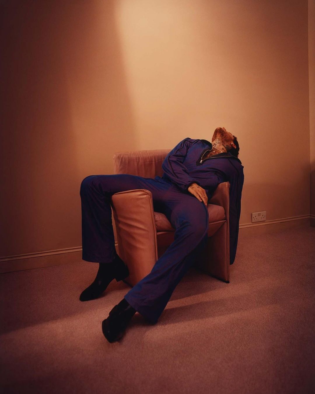 A person in a blue jumpsuit reclines in a pink armchair in a room with peach walls and carpet, lit by a dramatic light source.