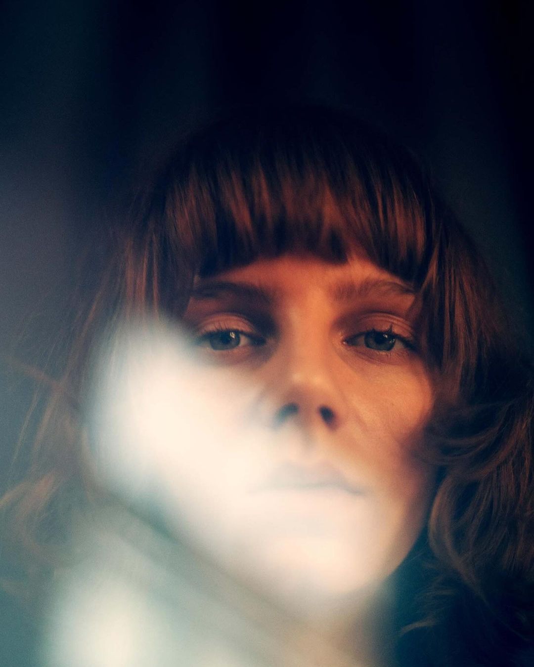 Close-up portrait of a woman with bangs looking into the camera with soft warm lighting and lens flare obscuring part of the frame.