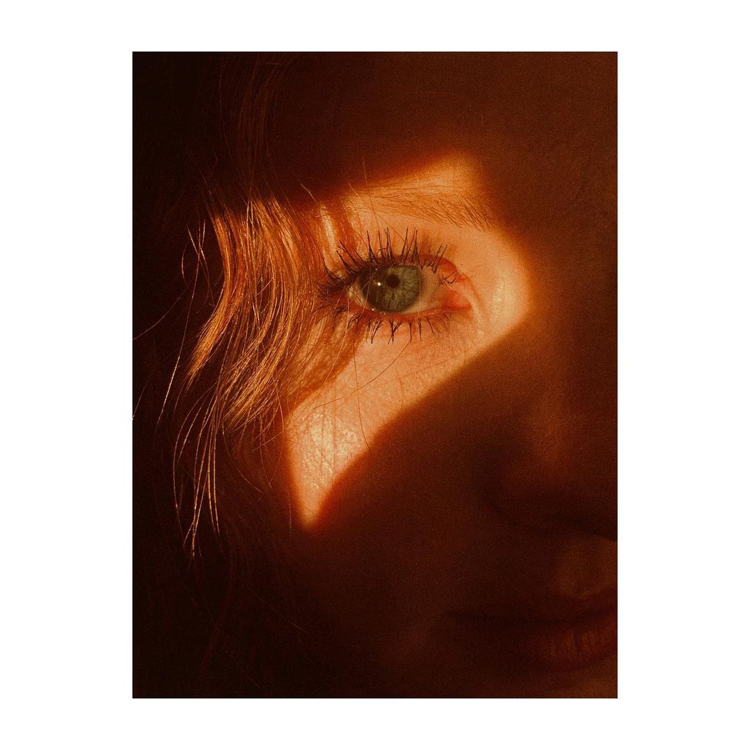 Close-up portrait of a woman's face, featuring her eye and the side of her face, partially shadowed with golden lighting.