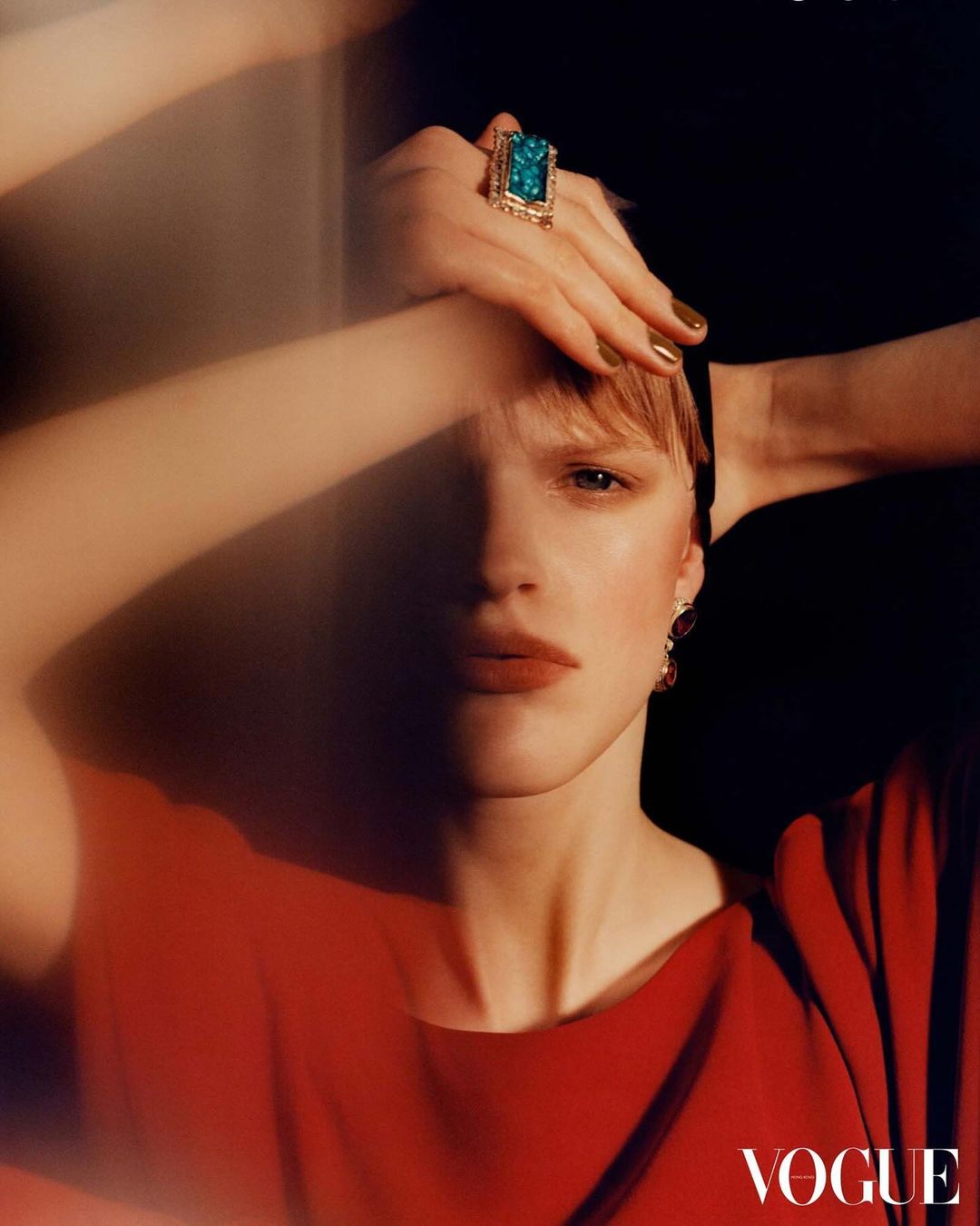 Close-up fashion shot of a woman in a red dress wearing a large ring, with hands above her head against a black background.
