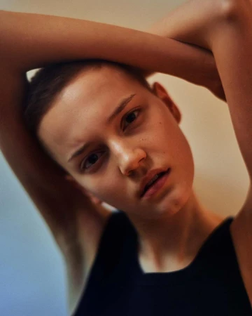 Close-up portrait of a person with short hair wearing a dark tank top with arms raised framing their head, photographed by Rory Payne.