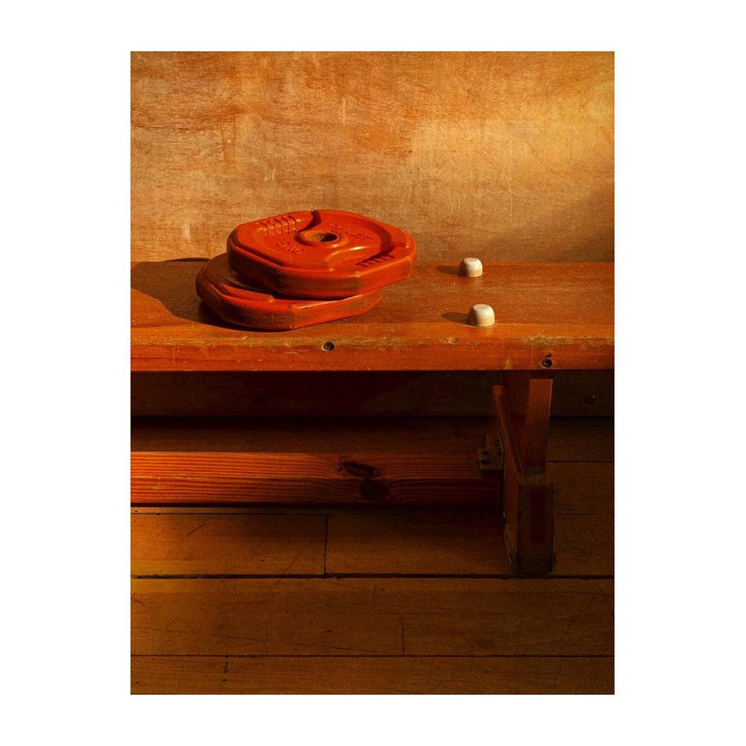 Two red weight plates are stacked on a worn wooden bench with two white cylindrical objects in a warm-toned indoor setting.