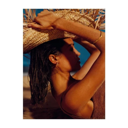 Close-up profile of a woman wearing a wide straw hat and brown knitted top against a sandy beach and blue ocean.
