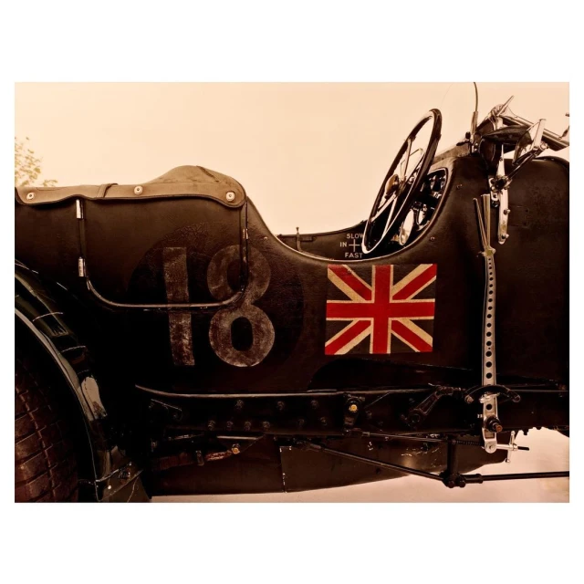 Vintage-style photograph of a classic race car featuring the number 18 and a Union Jack flag, set against a soft, neutral backdrop.
