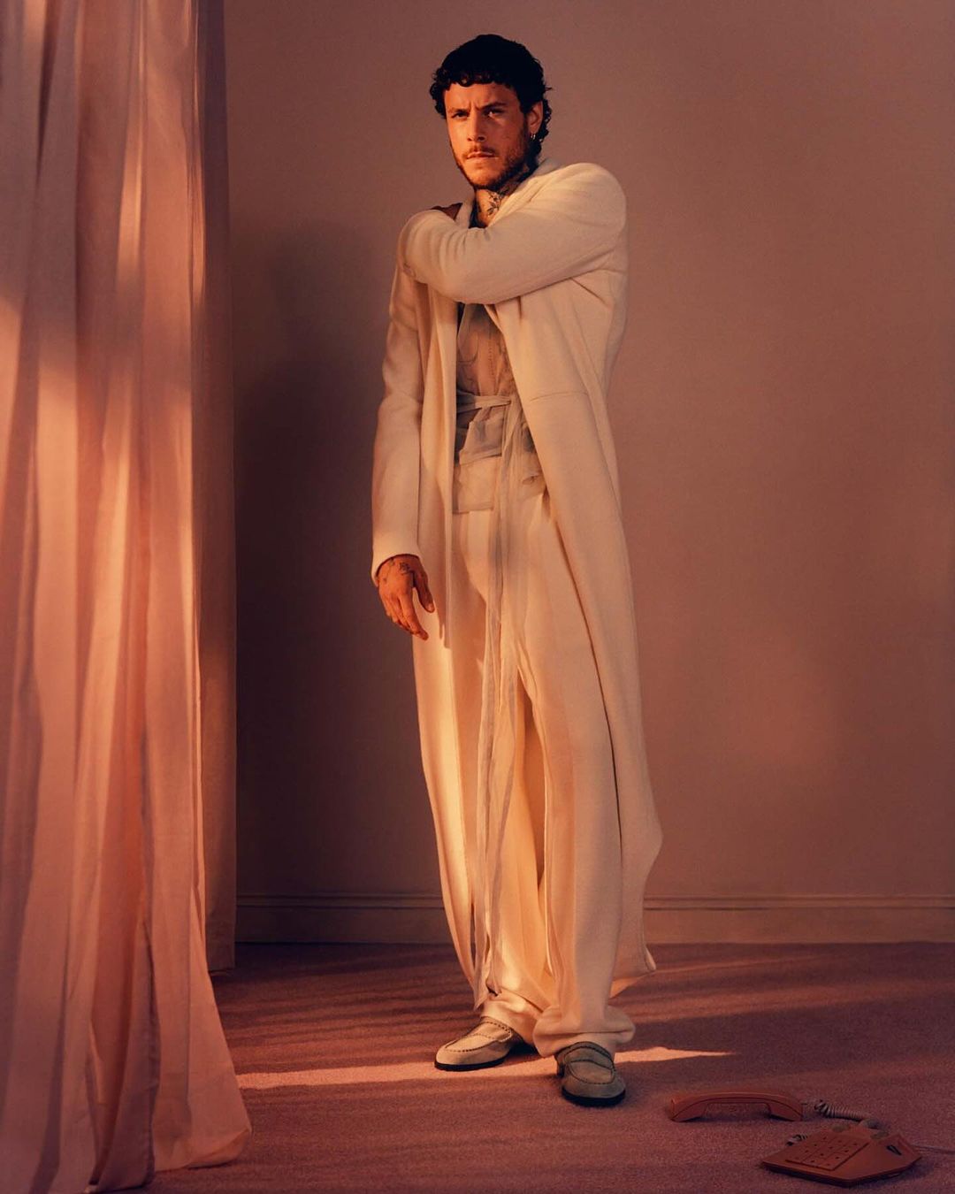 Portrait of an adult male in an off-white robe and pants, posing indoors against a plain backdrop, with soft light and shadows.