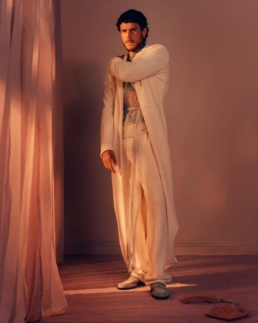Portrait of an adult male in an off-white robe and pants, posing indoors against a plain backdrop, with soft light and shadows.