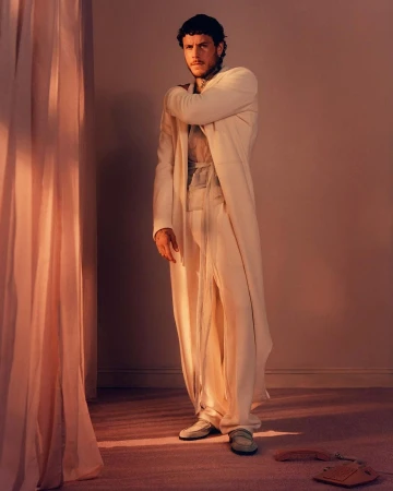Portrait of an adult male in an off-white robe and pants, posing indoors against a plain backdrop, with soft light and shadows.