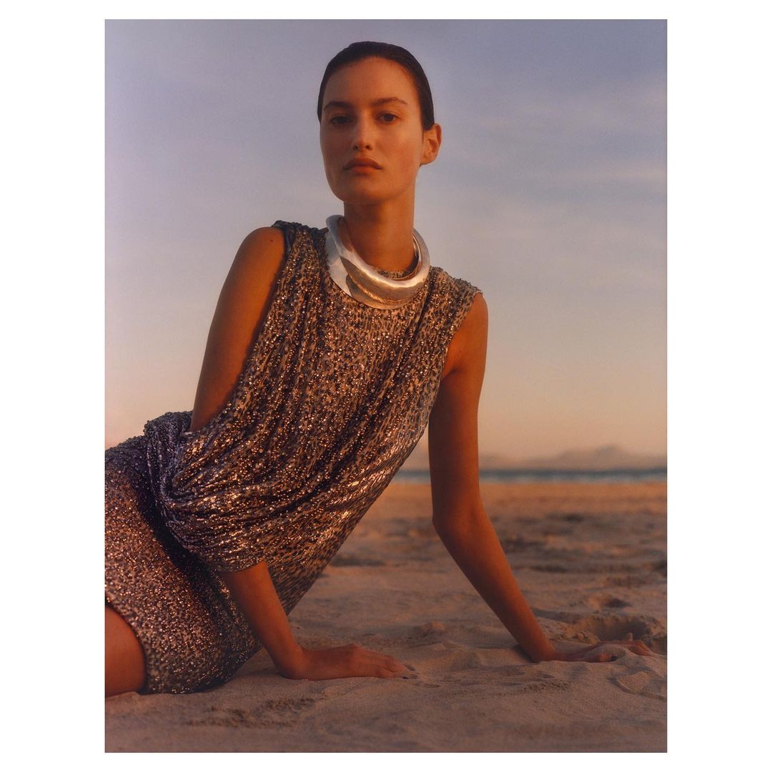 A woman poses in a sparkly dress and statement jewelry on a sandy beach at sunset with a distant ocean view, creating a fashionable, serene scene.