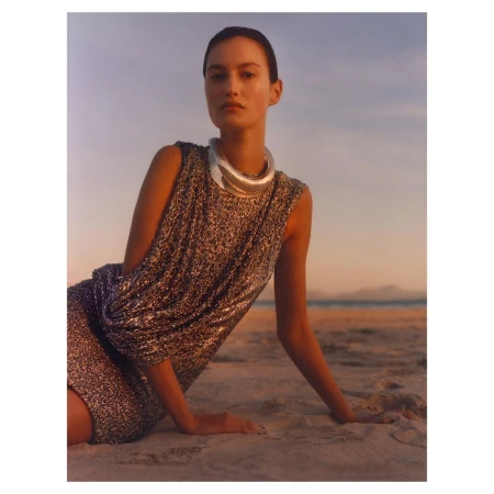 A woman poses in a sparkly dress and statement jewelry on a sandy beach at sunset with a distant ocean view, creating a fashionable, serene scene.
