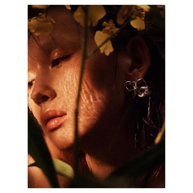 Close-up of a young adult woman with silver earrings, framed by yellow orchids and green leaves, with warm, diffused lighting.