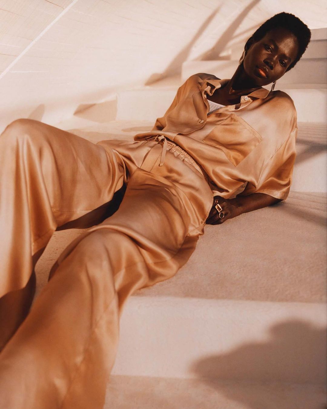 A dark-skinned woman in a light peach-colored satin shirt and pants poses on a white staircase in a fashion shot with natural lighting.