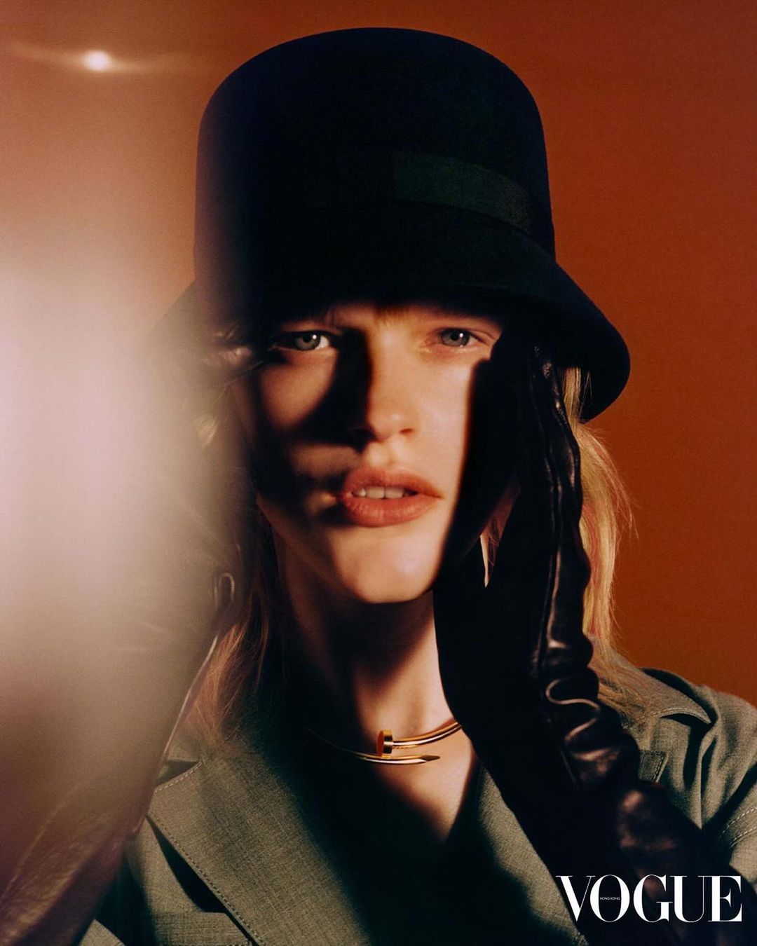 Fashionable woman in hat and gloves poses indoors for a Vogue magazine portrait with orange background and bright studio lighting.