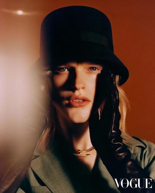 Fashionable woman in hat and gloves poses indoors for a Vogue magazine portrait with orange background and bright studio lighting.