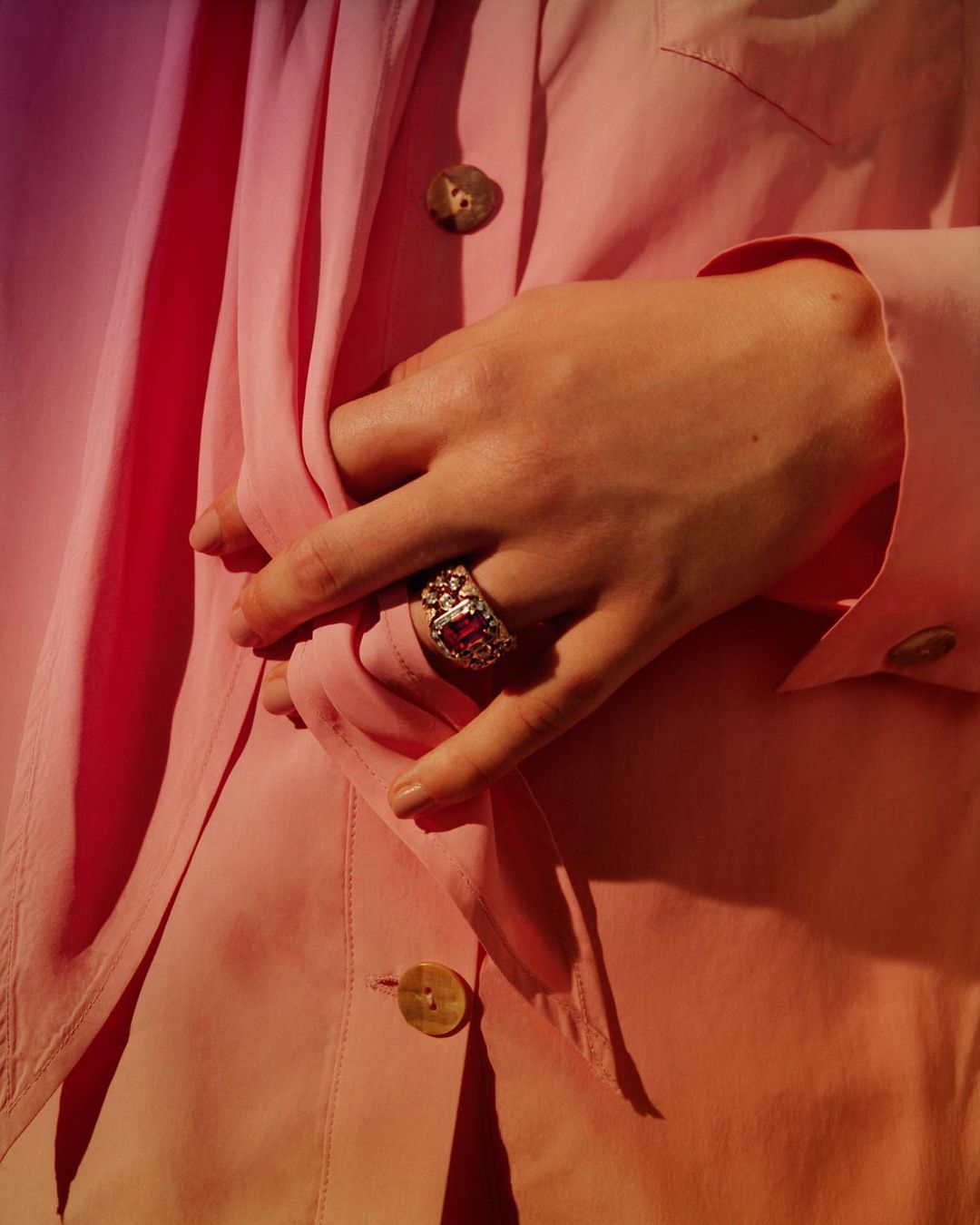 Close-up on an adult female's hand wearing a large ring with a pink gem, set against a light pink blazer with brown buttons.