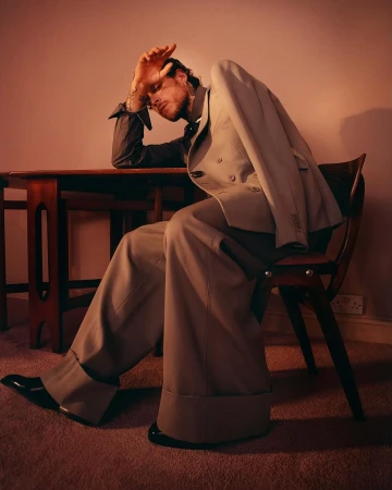 Man in a double-breasted suit sits at a wooden table and chair, his head resting on his hand in a studio portrait.