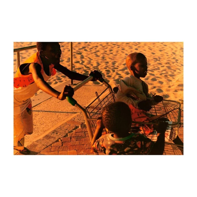 A dark-skinned adult woman pushes a metal shopping cart with two dark-skinned children as passengers across a sandy beach during the day.