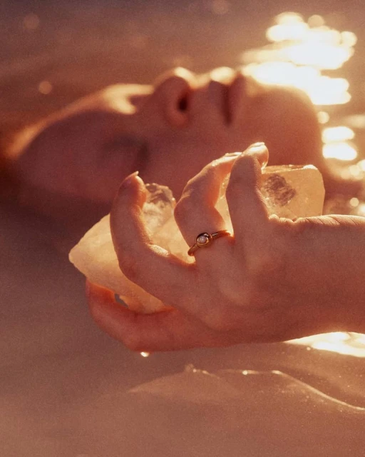 Close-up of a person in water holding a clear quartz crystal, with a gold ring on their finger, bathed in golden, soft lighting.