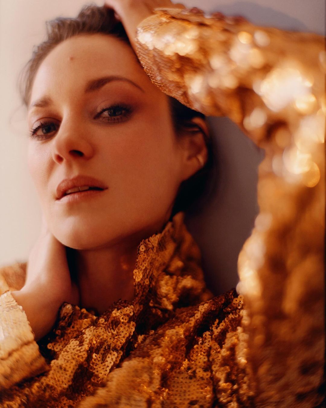 Close-up studio portrait of Marion Cotillard wearing a gold sequined jacket, fair skin, light makeup, eye contact, arms raised.