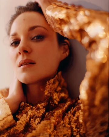 Close-up studio portrait of Marion Cotillard wearing a gold sequined jacket, fair skin, light makeup, eye contact, arms raised.