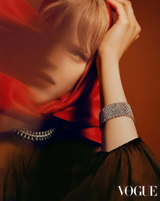 Fashion editorial shot of a model wearing a mesh bracelet and draped red scarf, posing against a brown backdrop with Vogue logo.