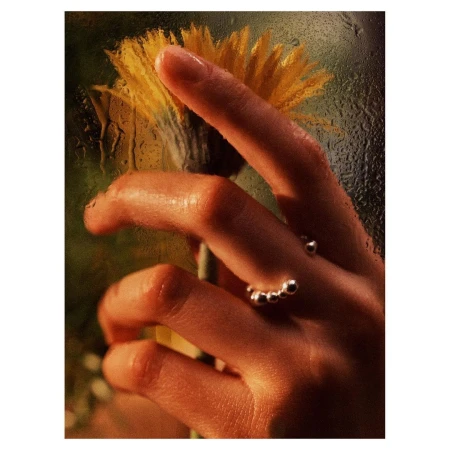 Close-up of a person's hand wearing a ring, holding a yellow flower against a window with water droplets, soft warm lighting.