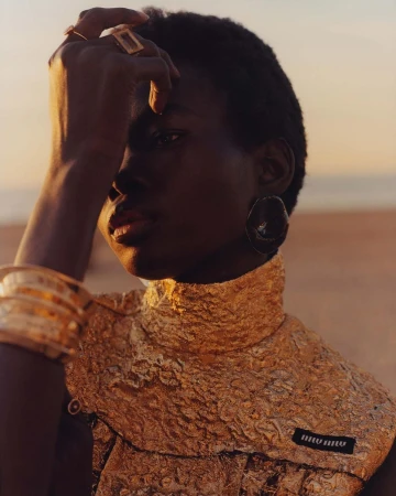 Fashion shot of a dark-skinned woman with gold jewelry and dress on a beach at sunset, featuring a close-up and warm color palette.