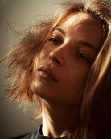 Close-up portrait of a woman with tousled hair, angled slightly upward with warm sunlight illuminating her face.