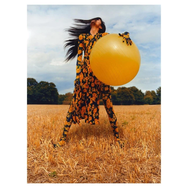 Fashion shot of a woman in a floral outfit holding a large yellow ball outdoors in a golden field under a cloudy sky.