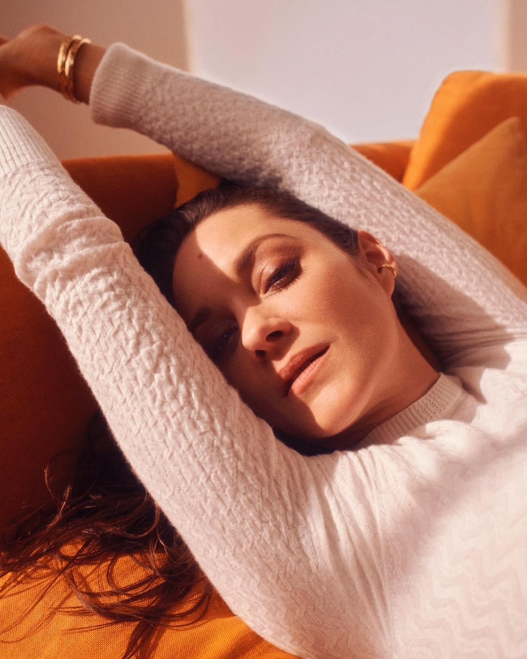 Woman with brown hair in a white sweater reclines on an orange couch, arms raised. Portrait orientation, warm lighting.