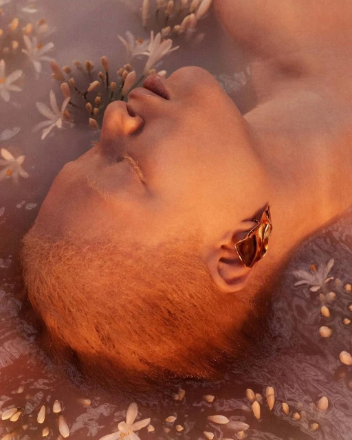 Close-up of a person in water surrounded by small white flowers, gold leaf ear cuff, and warm-toned studio lighting.