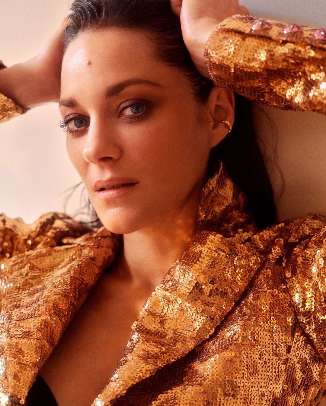Close-up portrait of a woman with dark hair wearing a gold sequined jacket, her hands near her head, looking at the camera indoors.