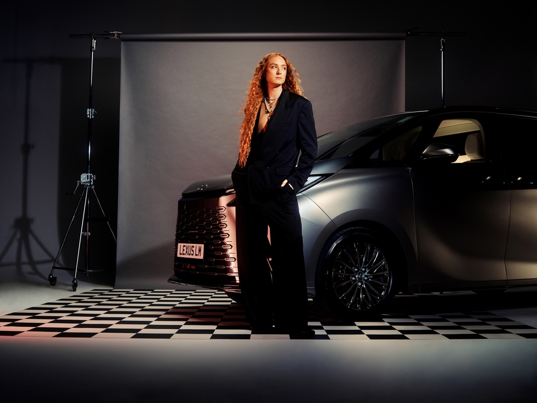 Stylish adult female model in a black suit poses next to a dark grey Lexus LM car in a photo studio setting with checkerboard floor.