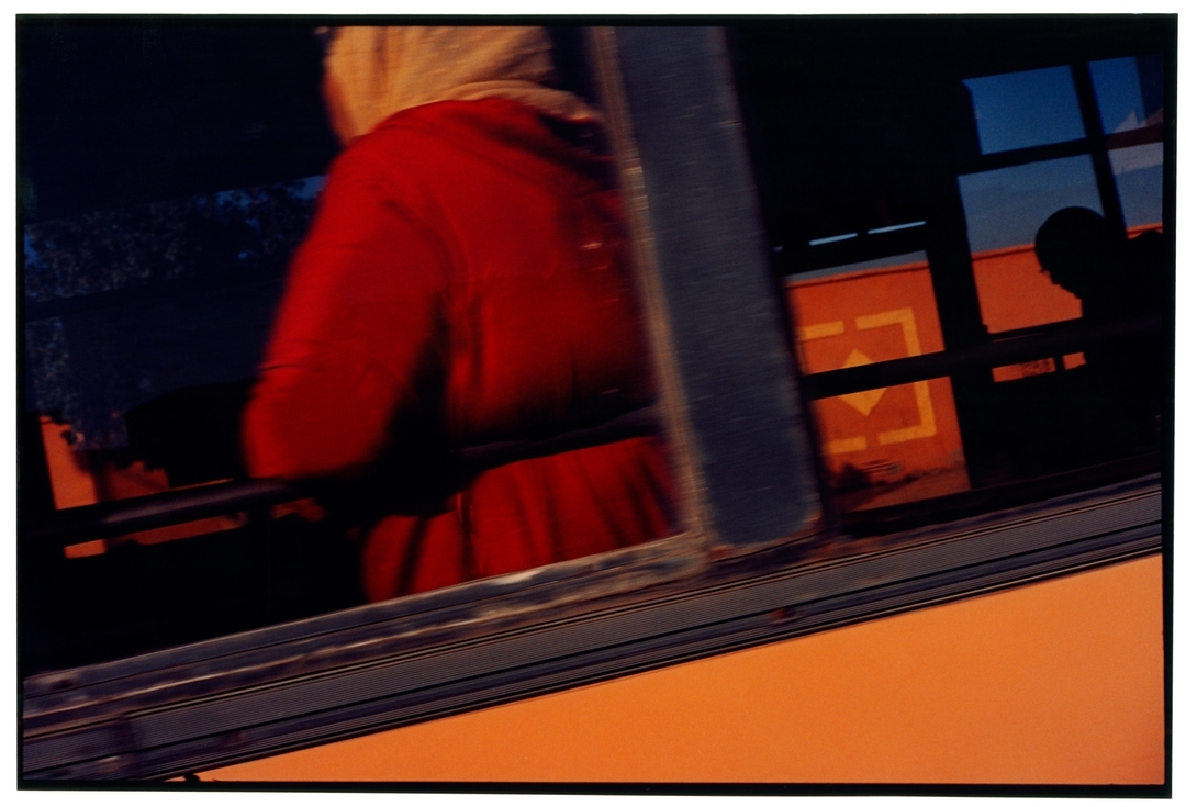 Abstracted image of a passenger in red in a bus, seen through a blurred window with an orange exterior and silhouetted head.