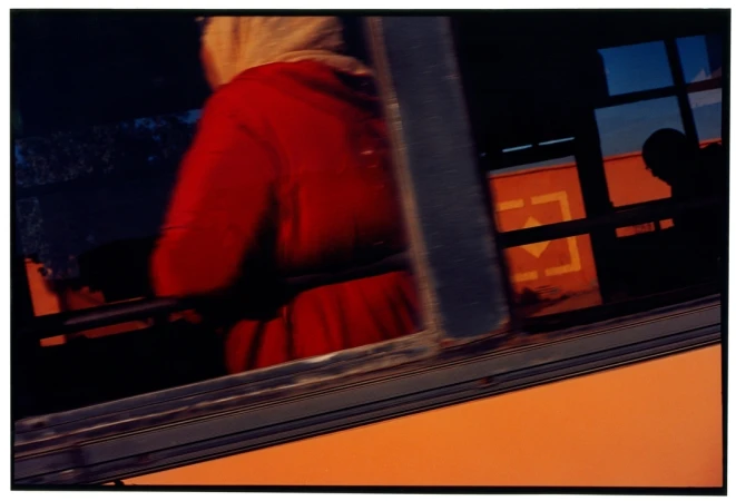 Abstracted image of a passenger in red in a bus, seen through a blurred window with an orange exterior and silhouetted head.