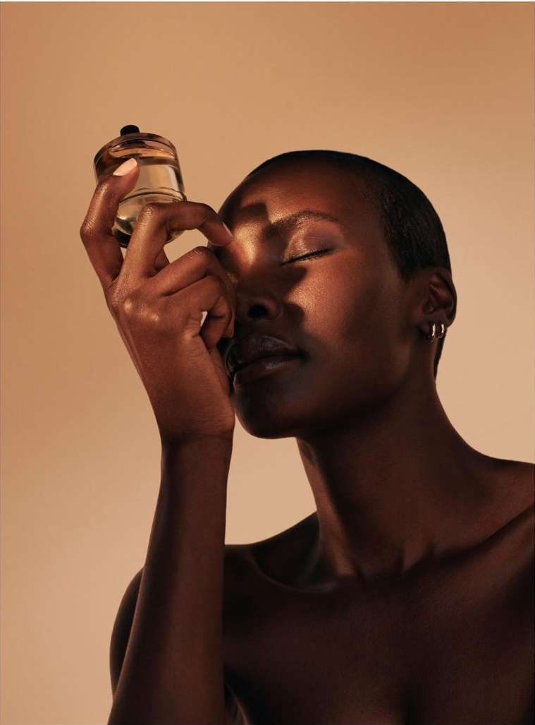 Striking beauty shot of a dark-skinned woman with closed eyes, holding a small glass container, set against a light brown background.