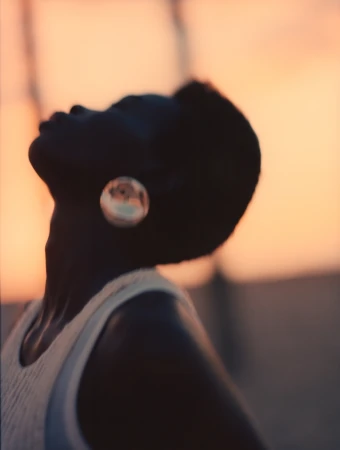 Profile portrait of a Black model in a ribbed cream tank top looking upward against a warm sunset sky background.