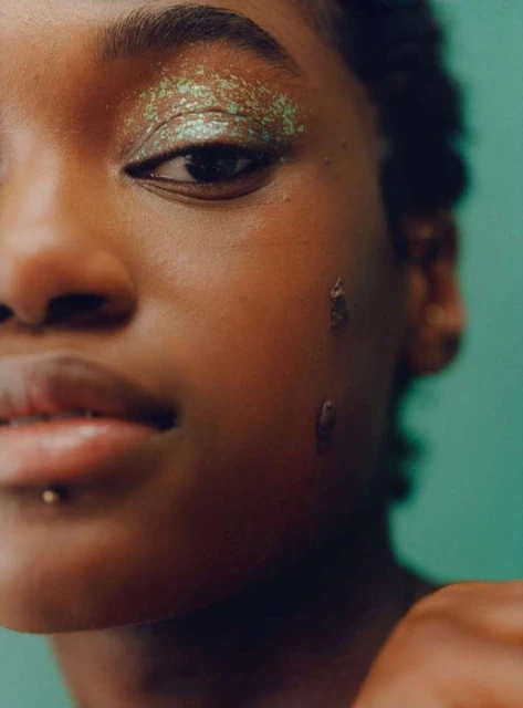 Close-up portrait of a woman with green glitter eye makeup and a lip piercing against a teal background, showcasing beauty and fashion.