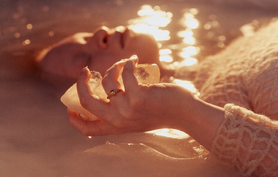 Close-up of an adult woman floating in water holding crystals, wearing a ring and textured top in warm lighting. Peaceful scene.