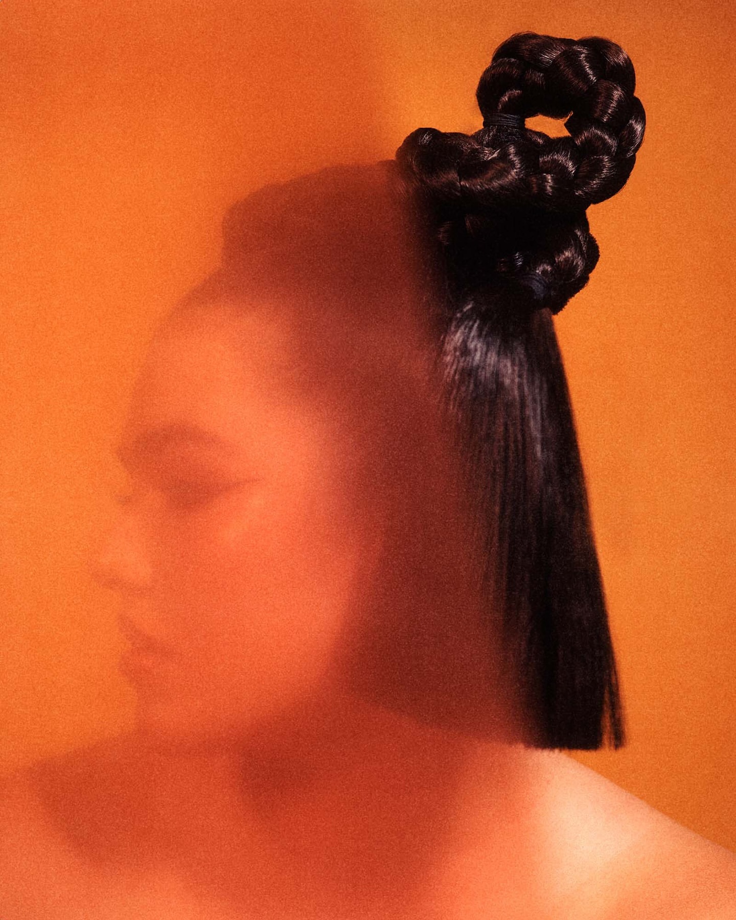 Profile portrait of a woman with sculpted black braided updo against warm orange background by Rory Payne