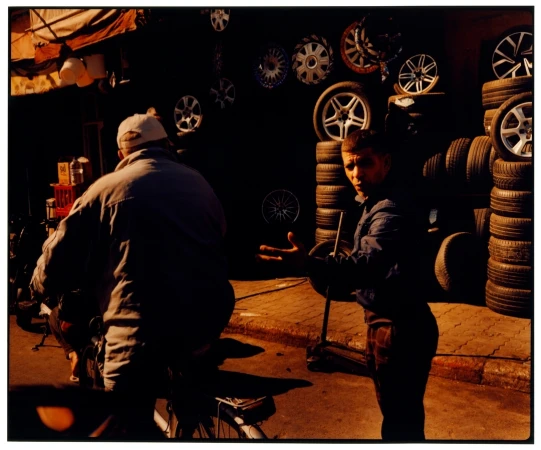 Two men are outside of a store, one riding a bicycle, while the other stands near stacked tires and rims for sale.