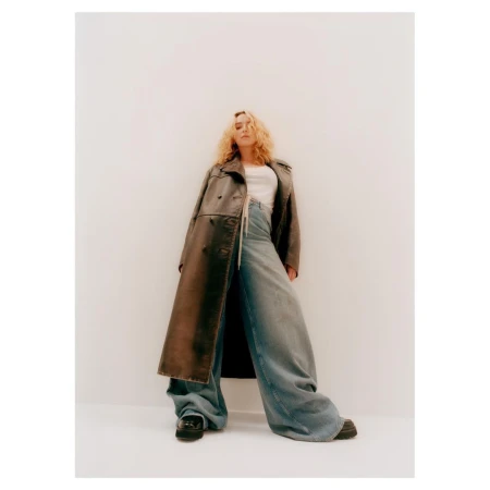 Woman with curly blonde hair wearing a long leather trench coat and wide-leg jeans against a plain white studio background.