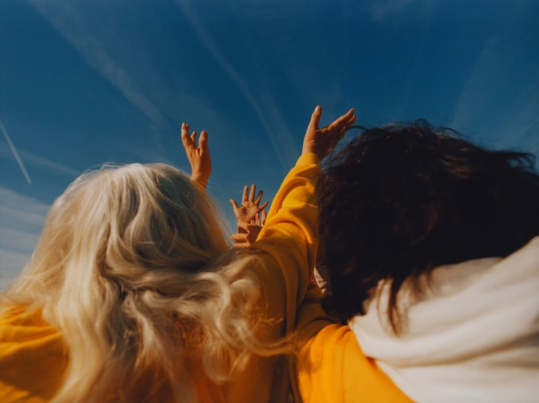 A group of three people are seen from behind in a low-angle shot under a bright blue sky, raising their hands upwards.