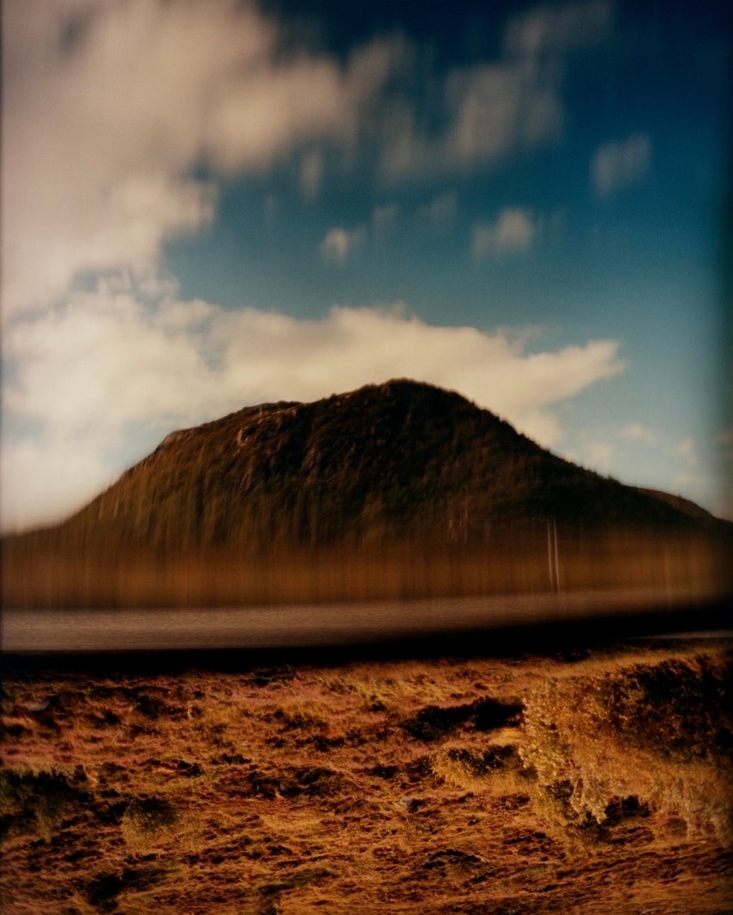 Wide shot of a grassy field landscape leading to a dark, rocky hill with a blue sky overhead with blurred, streaky white clouds.