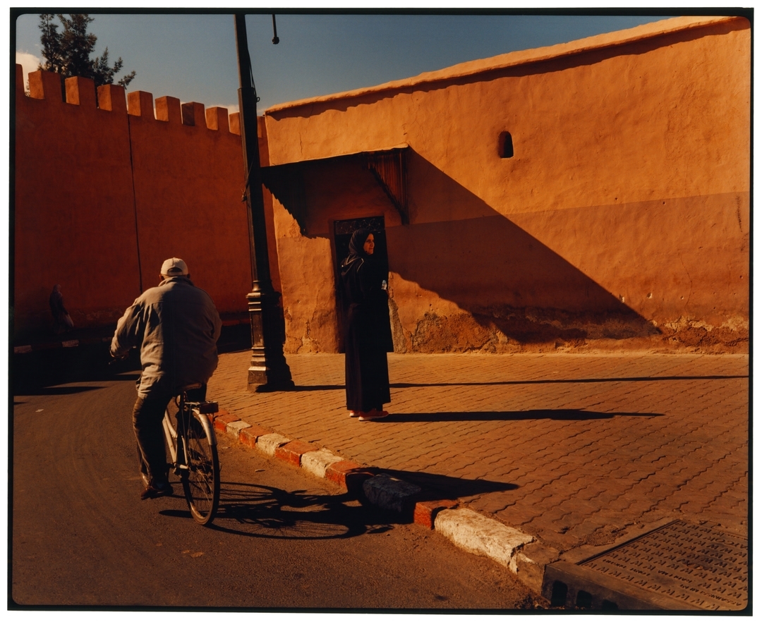 Warm-toned street scene featuring a man riding a bicycle and a woman in a hijab standing near a building, bathed in strong sunlight.