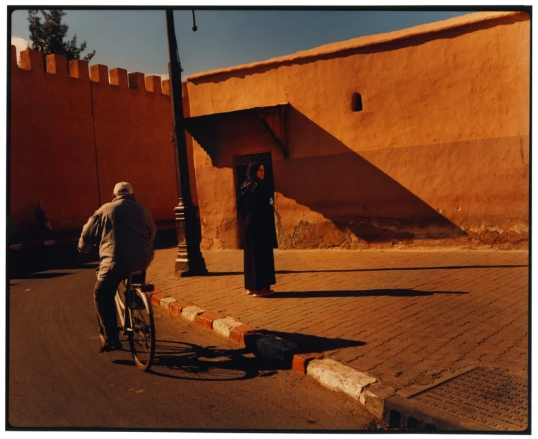 Warm-toned street scene featuring a man riding a bicycle and a woman in a hijab standing near a building, bathed in strong sunlight.