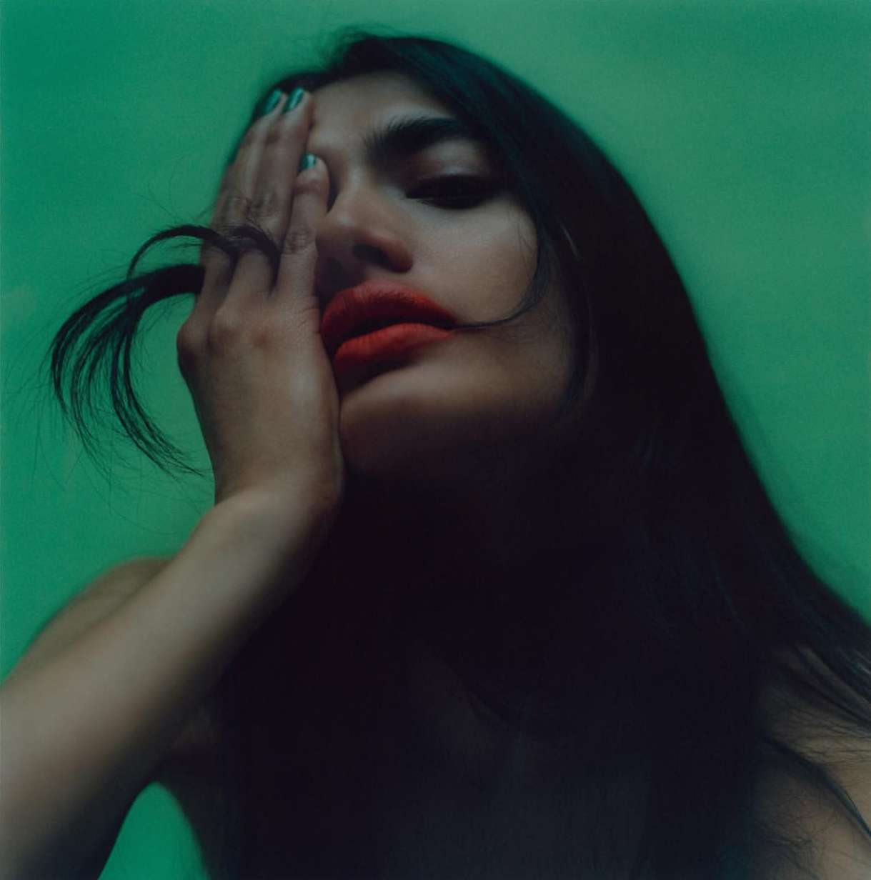Close-up portrait of a woman with long dark hair covering one eye with her hand against a solid green background.