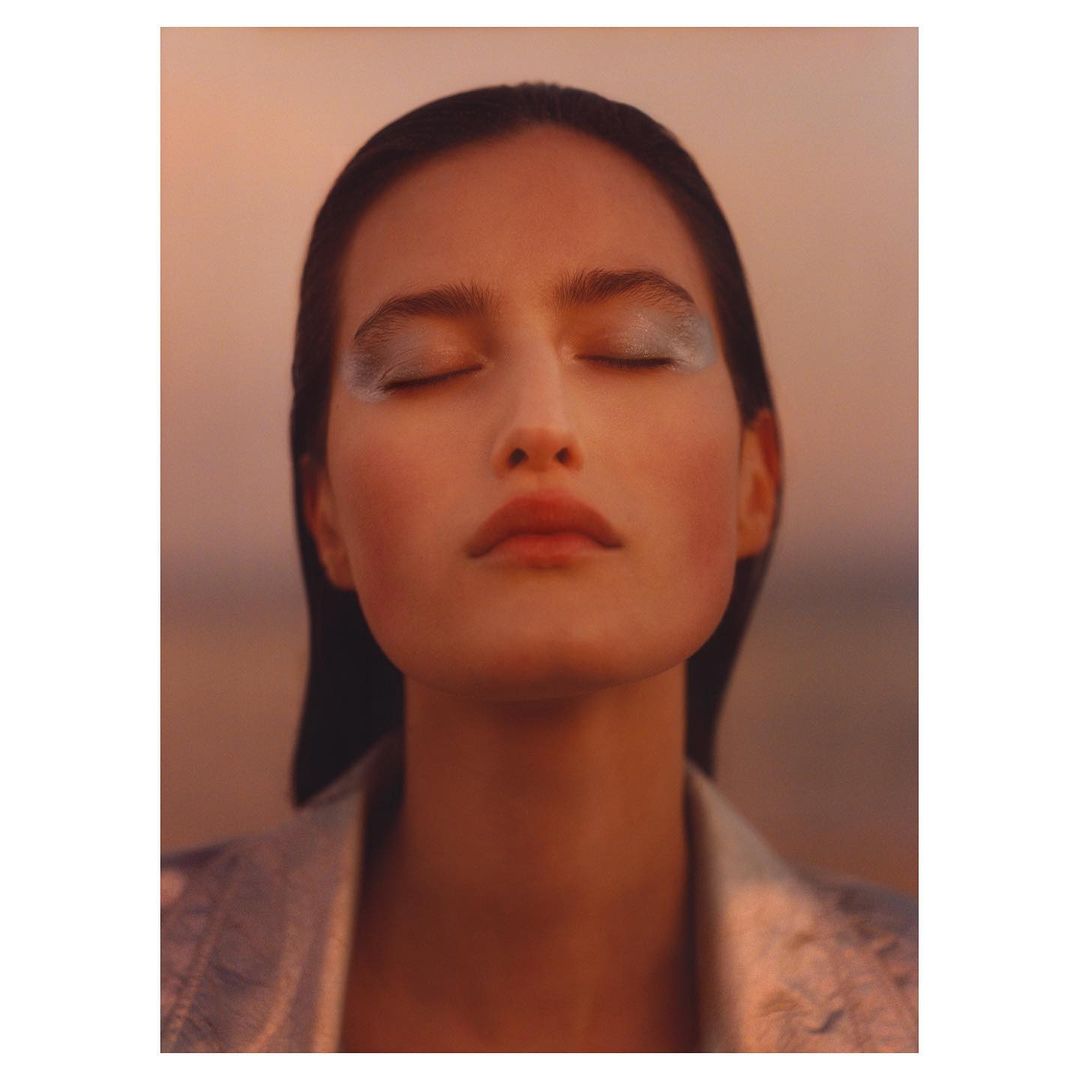 Close-up shot of an adult woman with closed eyes, wearing makeup and a silver jacket, with a blurred peach and grey background.