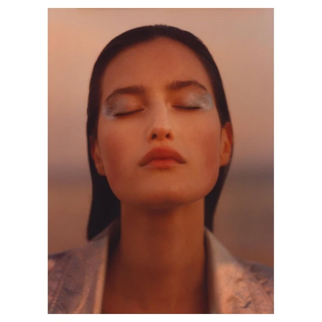 Close-up shot of an adult woman with closed eyes, wearing makeup and a silver jacket, with a blurred peach and grey background.